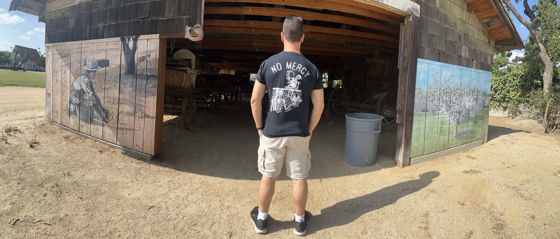 Man in "No Mercy" shirt standing outside a rustic barn with scenic paintings.