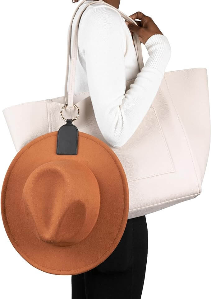 Person using magnetic hat holder clip to attach a brown hat to a large cream tote bag for hands-free convenience.