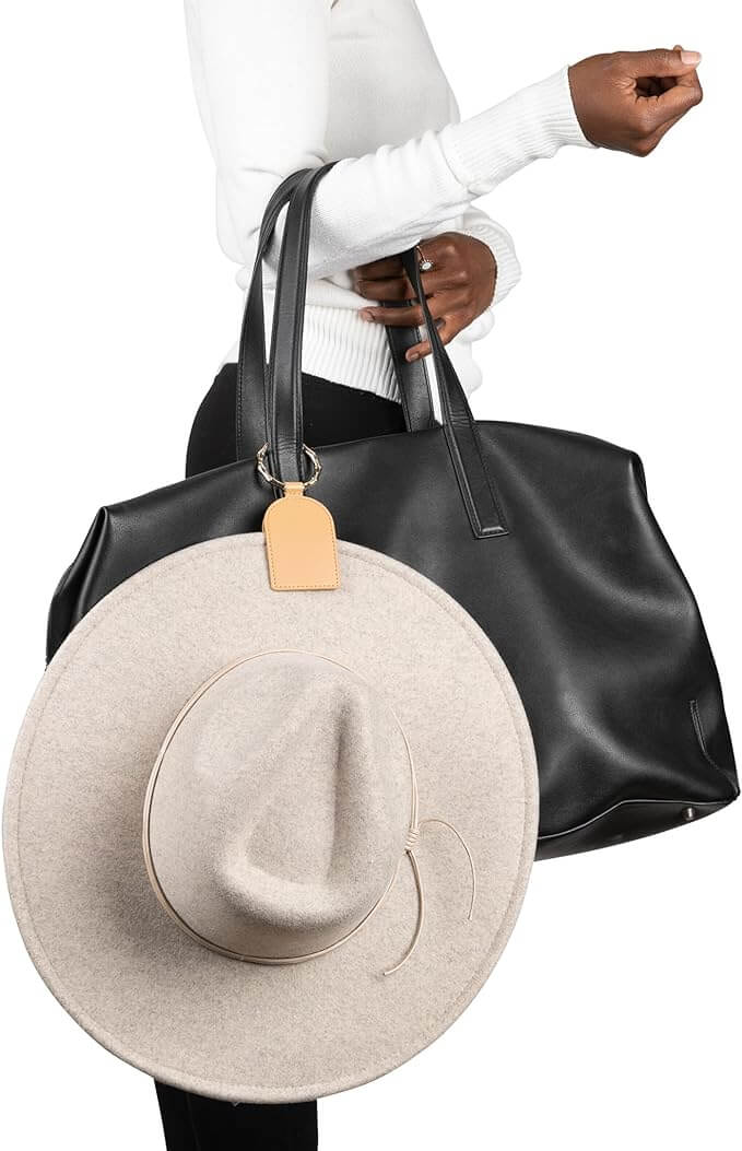 Woman holding a black bag with a tan DKMO Bell Hat Clip attached, securing a beige hat for convenient hands-free travel.