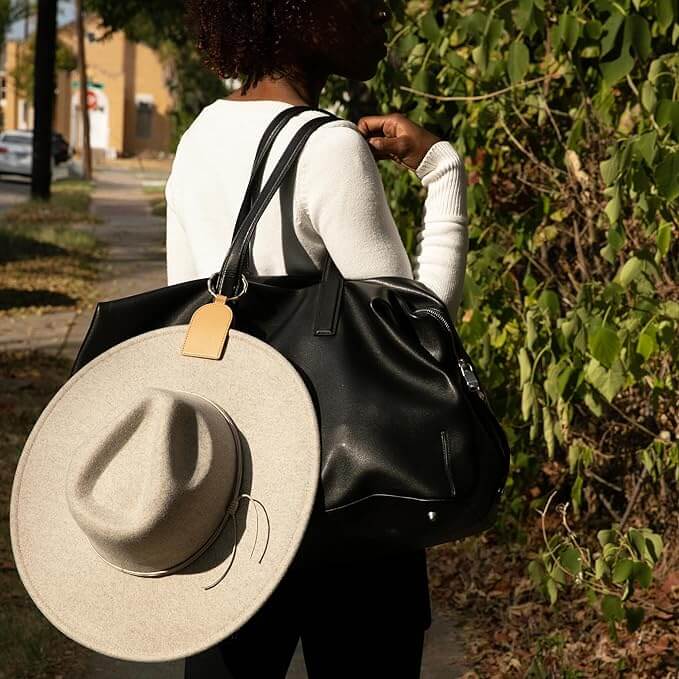 Woman carrying black bag with DKMO Bell Hat Clip holding a gray hat, walking outdoors.