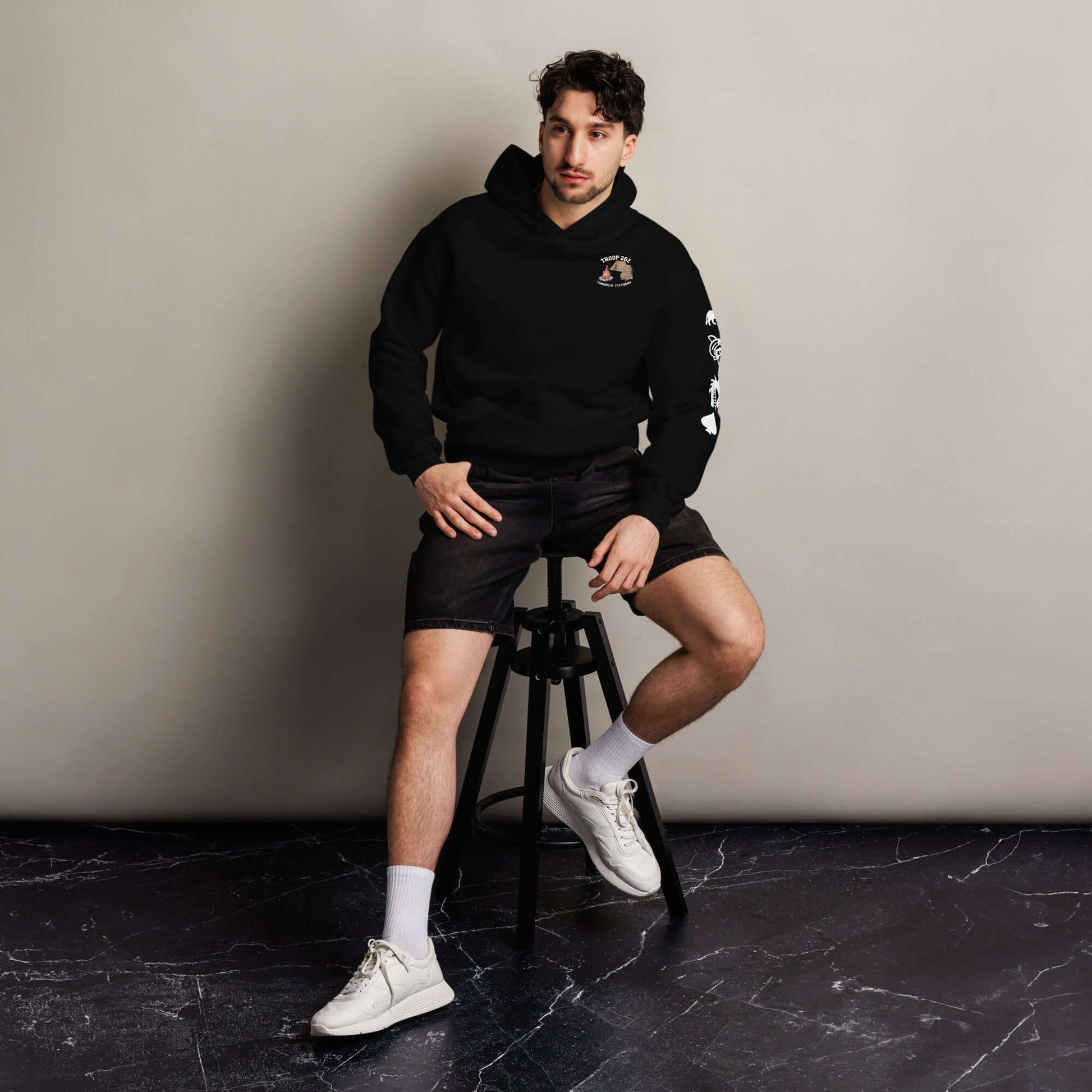 Man wearing a black oversized hoodie and shorts, sitting on a stool against a neutral background.